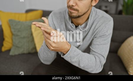 A focused young hispanic man with a beard examines his hands indoors, seated in a modern living room. Stock Photo
