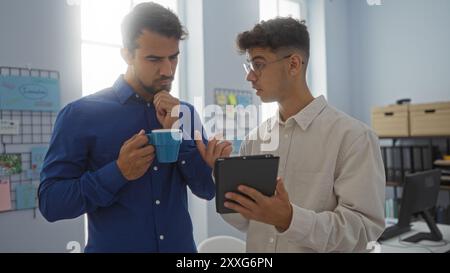 Two hispanic men discussing work in an office while one holds a mug and the other shows something on a tablet. Stock Photo