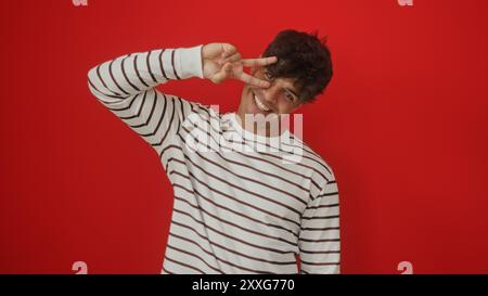 A handsome young hispanic man in a striped shirt poses with a cheerful smile against a vibrant red background, showcasing his playful personality. Stock Photo
