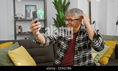 A cheerful senior man with glasses takes a selfie in a modern living room, showcasing technology and lifestyle. Stock Photo