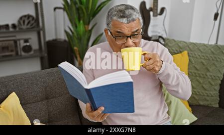 A senior man with glasses enjoys reading a book and drinking from a yellow mug in a cozy living room. Stock Photo