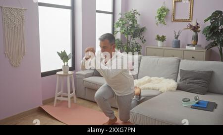 Grey-haired hispanic man practicing yoga on a mat in a cozy living room filled with plants and soft furnishings. Stock Photo