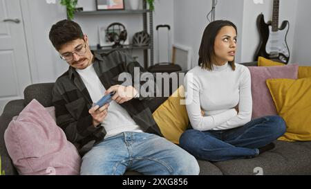 A man and woman sit on a couch, the man focused on his smartphone while the woman appears annoyed, in a modern living room. Stock Photo