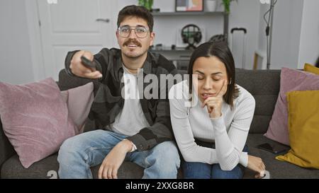 A bored woman and an engaged man sitting together on a couch with a remote control in a modern living room. Stock Photo
