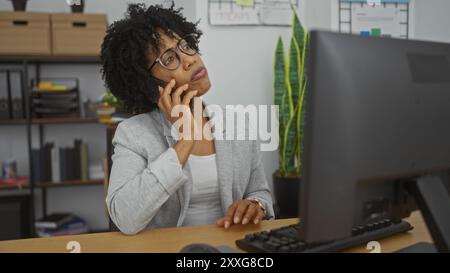 A beautiful young african american woman with curly hair works indoors at an office, talking on the phone, wearing glasses, and appearing thoughtful. Stock Photo