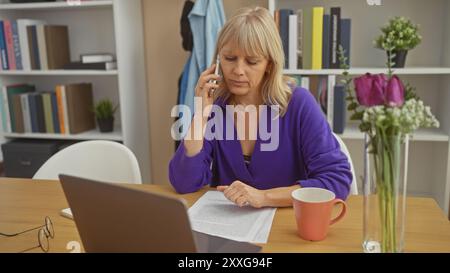 A mature woman engages in a serious phone conversation at home, surrounded by flowers and books. Stock Photo