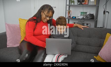 A smiling woman and boy enjoy using a laptop together on a cozy couch in a modern living room. Stock Photo