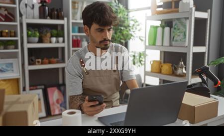 Young hispanic man with a beard working in a home decor store, using a laptop and smartphone, surrounded by various decorative items on shelves. Stock Photo