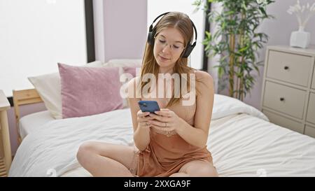 A young adult woman in a bedroom, wearing headphones and using a smartphone, depicts a casual, cozy lifestyle. Stock Photo