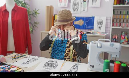A man in a hat rubs his eyes in frustration in a colorful tailor shop with sewing machine and fabric. Stock Photo