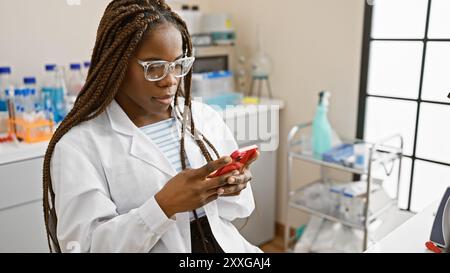 African american female scientist with braids using smartphone in laboratory Stock Photo