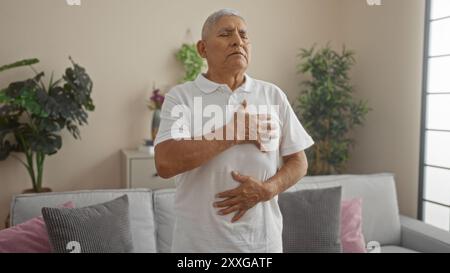 An elderly hispanic man with grey hair practicing deep breaths in his living room, standing with hands on chest and stomach, surrounded by plants and Stock Photo