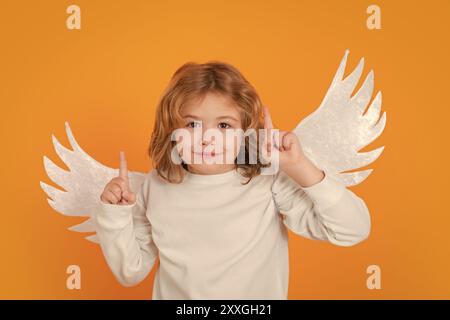 Child angel pointing up, point gesture, idea. Valentine's day. Blonde cute child with angel wings on a yellow studio background. Happy angel child. Stock Photo