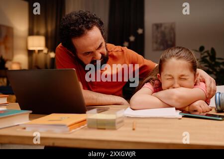 Father encouraging daughter while assisting with schoolwork in a cozy room, with books and laptop on wooden desk creating a warm study environment Stock Photo
