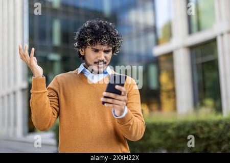 Man with curly hair standing outside modern office building looks at phone with frustration. Wearing a brown sweater, he expresses confusion or dissatisfaction with technology issues. Stock Photo
