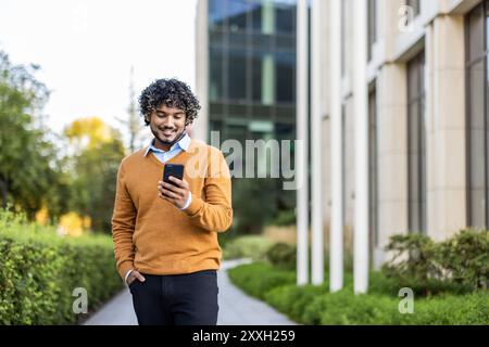 Young man in orange sweater uses phone while walking in urban area. Happy with technology interaction. Sunny day highlights blend of nature and architecture in modern city setting. Stock Photo