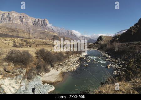 A mountain river in a mountainous rocky area near which there are power lines on the background of epic rocks and a blue sky. Concept of energy supply Stock Photo