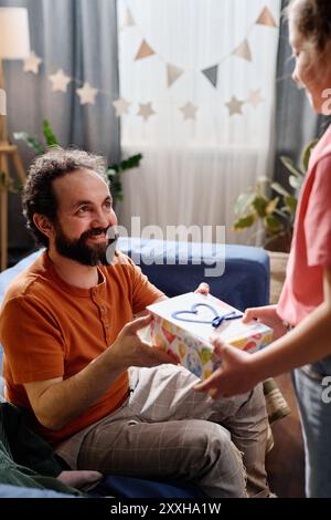 Man receiving a present from child while smiling and sitting on couch with festive decorations in background including star-shaped garlands and plants Stock Photo