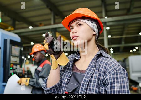 Engineers wearing safety helmets and gloves working on machinery in spacious industrial setting with focus on female engineer using radio communication Stock Photo