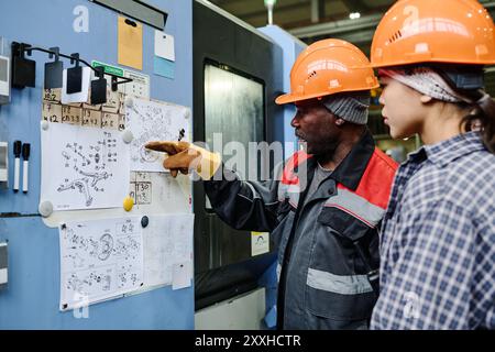 Two factory workers examining detailed blueprints pinned on a corkboard discussing important aspects of the technical drawings in an industrial setting Stock Photo