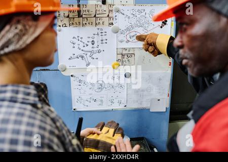 Two engineers reviewing detailed schematics on bulletin board, one pointing at specific part of the drawing while the other listens attentively wearing safety gear Stock Photo
