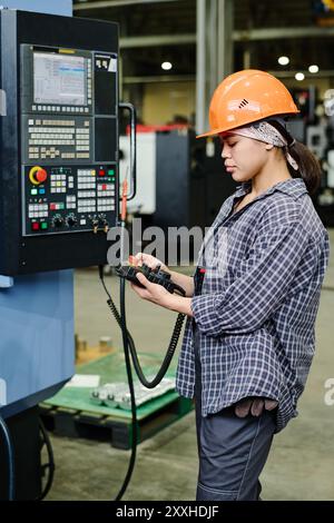 Worker in factory operating industrial machine using control panel wearing safety equipment including hard hat focusing on task in industrial setting Stock Photo