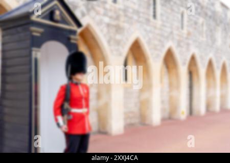 A blurred image of a British guard in a red uniform and a black bear skin hat standing in front of a stone building. Stock Photo