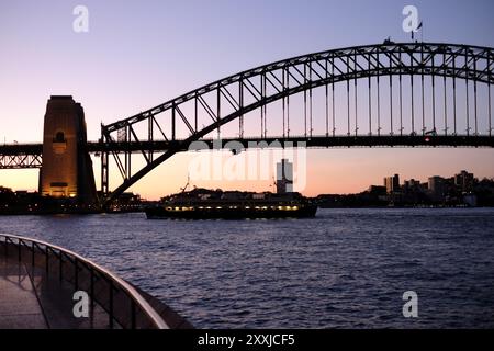 Sydney, Australia, the Art Deco design Sydney Harbour Bridge in silhouette at dusk with the sun setting behind the southern pylon as a ferry passes by. Stock Photo