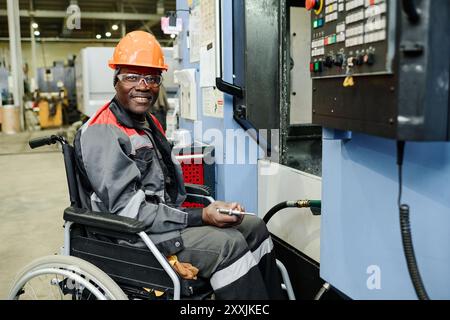 Industrial worker in wheelchair smiling while operating machinery in factory setting, showing determination and competence. Wearing helmet and safety gear, focused on task Stock Photo