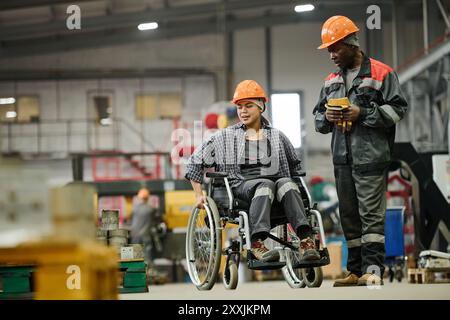 Wheelchair-using worker conversing with a colleague in an industrial warehouse setting, both wearing protective gear and helmets in their working environment, showcasing inclusivity and teamwork Stock Photo