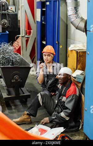 Workers engaged in communication on a busy industrial worksite with various equipment and tools visible. Safety gear including hard hats and protective clothing worn by workers present Stock Photo