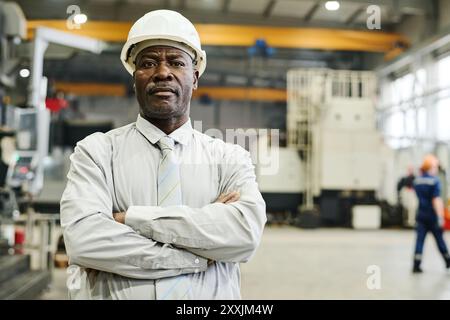 Portrait of African American engineer wearing white hardhat standing in factory with arms crossed, background showing industrial equipment and workers Stock Photo