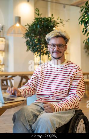 Portrait disabled man freelancer on wheelchair sits on adaptive place in coffee shop with laptop. Stock Photo