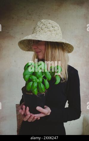 Vertical waist up portrait of a woman in beige summer hat holding a pot with basil Stock Photo