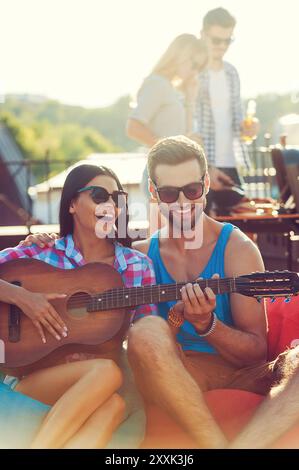 Guitar fun. Joyful young man teaching his girlfriend to play the guitar while two people barbecuing in the background Stock Photo