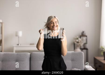 Cheery woman dancing in living room with smartphone in hand Stock Photo