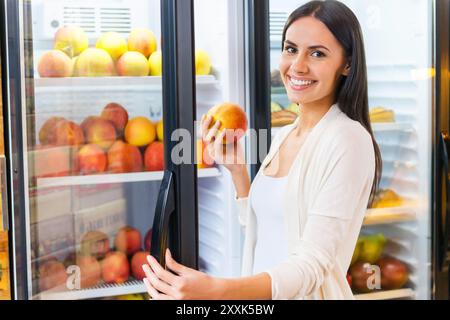 Choosing the freshest apple. Beautiful young smiling woman choosing apples from refrigerator in grocery store Stock Photo