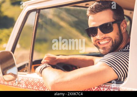 Enjoying his road trip. Cheerful young man smiling at camera and holding hand on steering wheel while sitting inside of his minivan Stock Photo