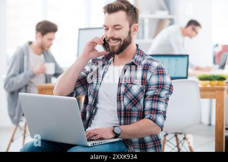 When the work is in full swing. Happy young man working on laptop and talking on the mobile phone while two people working in the background Stock Photo