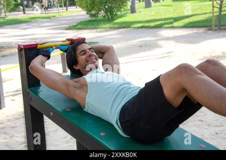A man doing leg exercises on an abdominal bench in a public park. He is wearing a green T-shirt and black pants. Stock Photo