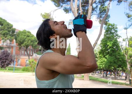 A man is drinking water from a bottle while wearing gloves. The scene is set in a park with trees and a bench. The man is taking a break from his work Stock Photo