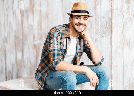 Casually handsome. Handsome young man wearing hat and looking at camera while sitting against the wooden wall Stock Photo