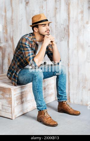 Deep in his thoughts. Handsome young man wearing hat and looking away while sitting against the wooden wall Stock Photo