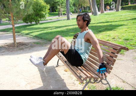 A man doing leg exercises on a bench in a public park. He is wearing a green tank top and black pants. Stock Photo
