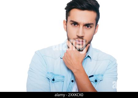 Thinking about solution. Thoughtful young Indian man holding hand on chin and looking at camera while standing against white background Stock Photo