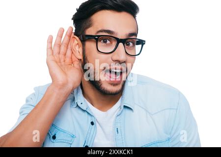 What did you say? Curious young Indian man holing hand near ear and looking at camera while standing against white background Stock Photo
