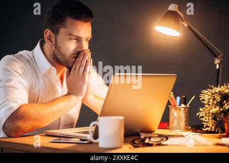 Thinking about solution. Thoughtful young man holding hands clasped near face while sitting at his working place at night time Stock Photo