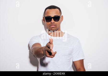 Hands up! Serious young African man in sunglasses gesturing finger gun while stretching out hand and standing against white background Stock Photo