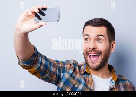 Cheerful selfie. Cheerful young man in shirt holding mobile phone and making photo of himself while standing against grey background Stock Photo