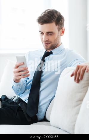 Feeling comfortable while doing business. Smiling young businessman holding mobile phone while sitting on sofa Stock Photo